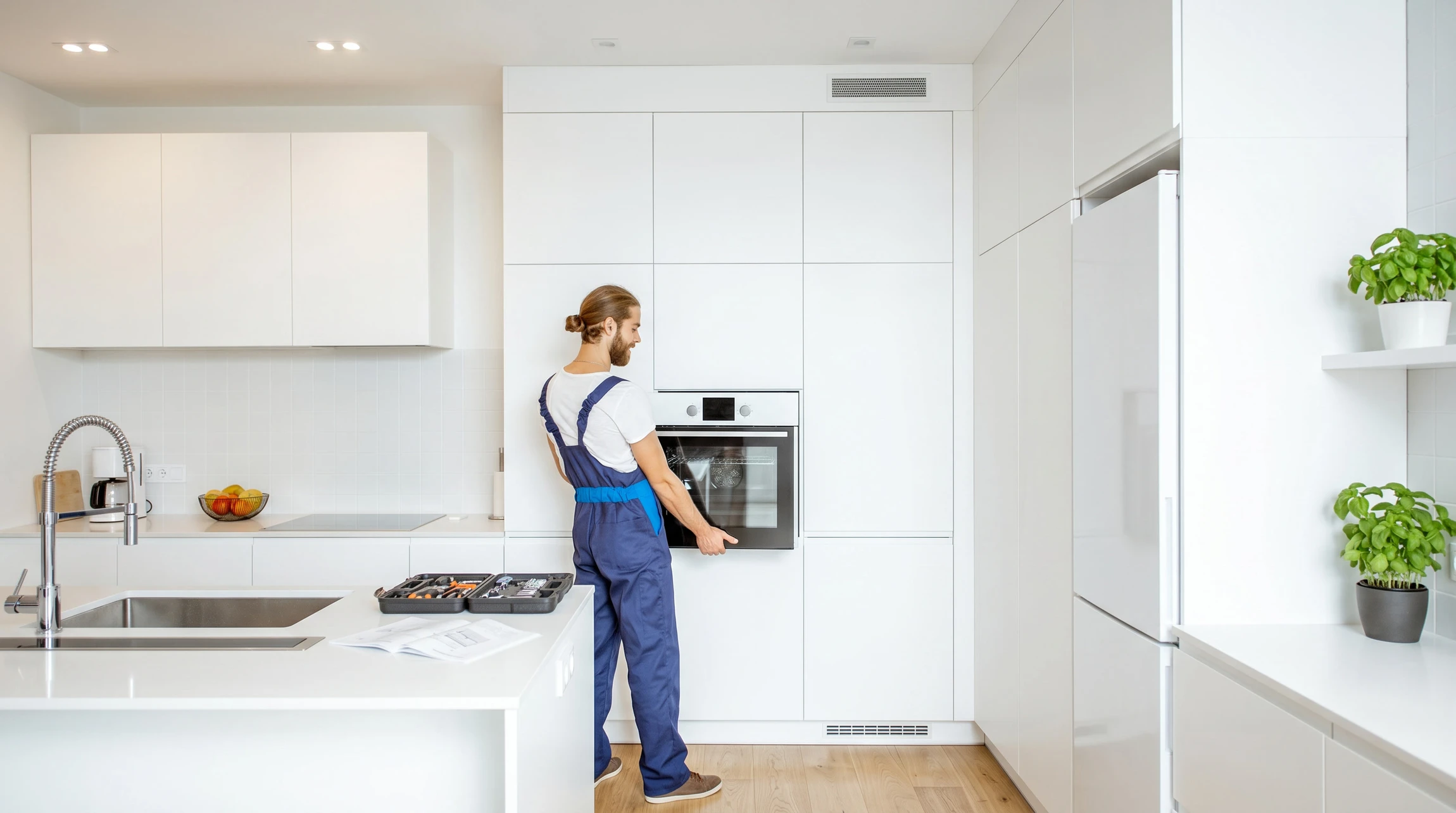 Technicien en combinaison bleue et pull blanc effectue une intervention de maintenance sur un appareil électroménager blanc dans une cuisine moderne